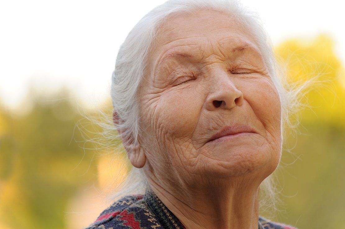 Eine Seniorin hält ihr Gesicht in die Sonne, die Augen sind geschlossen und sie lächelt.
