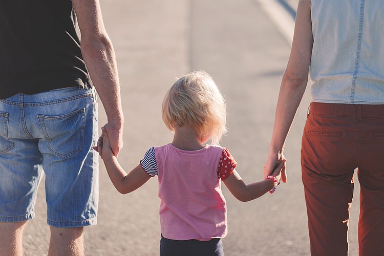 Ein kleines Mädchen hält rechts die Mutter und links den Vater an der Hand